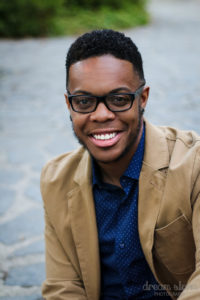 photo of Kyle, smiling in glasses, a snappy tan blazer and a navy blue button down with a small white pattern