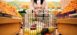 person with long hair and a light grey sweater pushing a shopping cart through a produce isle lined with citrus