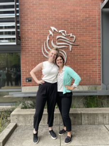 Kelsey and Emily, wearing white tops and black slacks, standing in front of Pacific U's qilin mascot in metal wall art on a brick wall
