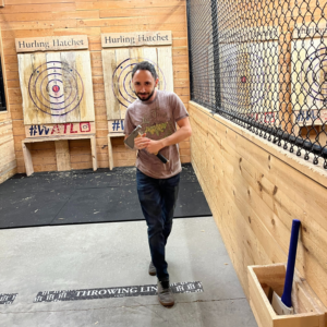 andrew, holding an axe in an axe throwing range, wearing a taupe shirt and black pants.