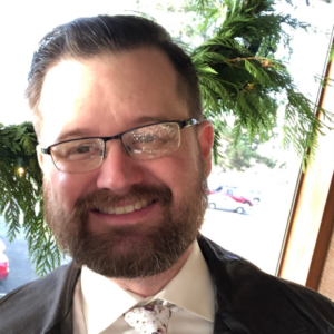 Randall, smiling with glasses. Wearing a dark jacket, a floral tie, a white button down, and glasses.