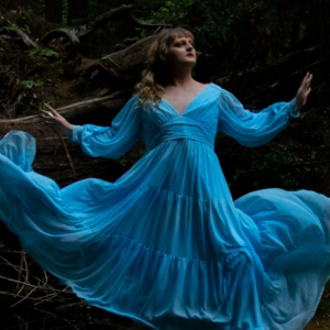 Melrose in a pose with arms out, wearing a long, flowing blue dress. Her long blonde hair is swept to one side. Dark black background.