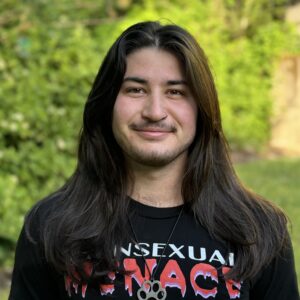 Alex, wearing a black shirt with white and pink text on the front. Trees in the background.